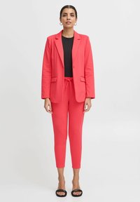 Korálový blejzr a odpovídající slim-fit kalhoty vyrobené z elastického materiálu; mají klopu, knoflíkové zapínání a stahovací pas. Černý top a sandály.