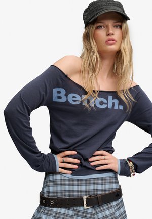 Ung kvinde iført en marineblå off-shoulder top med teksten "Bench.", ternet nederdel, sort bælte, sort kasket og flere armbånd, hænderne på hofterne.