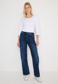 Wijdvallende donkerblauwe denim jeans met een vervaagde textuur en witte stiksels. Gecombineerd met een wit longsleeve shirt en witte sneakers.