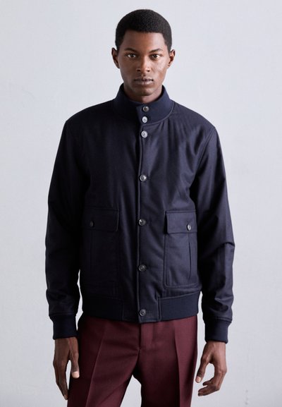 Giacca blu navy con colletto alto, chiusura frontale con bottoni e due tasche anteriori; realizzata in tessuto liscio con polsini e orlo a coste.