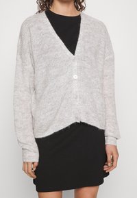 Lichtgrijze gebreide cardigan met een V-hals, vier knopen, verlaagde schouders en een verkorte zoom; gedragen over een zwarte jurk.