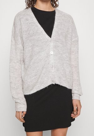 Cardigan en tricot gris clair avec un col en V, quatre boutons, épaules tombantes et un ourlet court ; porté sur une robe noire.