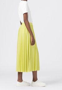 Personne portant un haut blanc à manches courtes, une jupe midi plissée vert clair et des baskets blanches se tenant de côté sur un fond blanc.