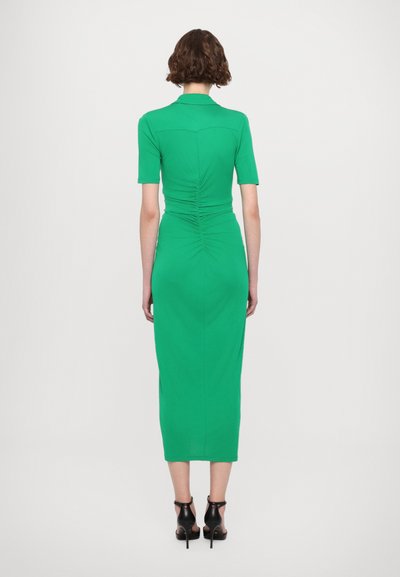 Vestido verde ajustado con mangas cortas, detalles fruncidos en la parte trasera y longitud hasta la mitad de la pantorrilla. Combinado con zapatos de tacón alto negros.