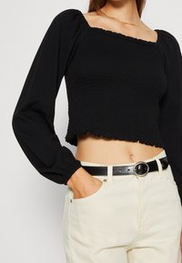 Top cropped nero con scollatura quadrata, texture smockata, maniche lunghe a sbuffo e orlo arricciato, abbinato a jeans a vita alta color crema e una cintura nera.