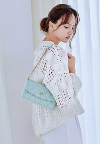 Sac à main matelassé vert menthe avec des accents en perles et des accessoires en or, porté en bandoulière. Le modèle porte un pull en maille blanche avec des motifs ajourés.
