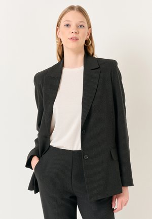 Blazer nero a righe sottili con revers a punta, chiusura a un bottone e tasche frontali. Indossato su una camicia color crema. Vestibilità sartoriale.