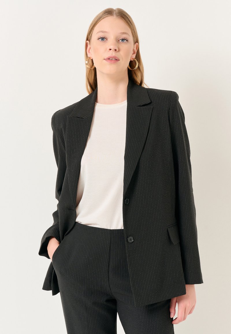 Blazer nero a righe sottili con revers a punta, chiusura a un bottone e tasche frontali. Indossato su una camicia color crema. Vestibilità sartoriale.