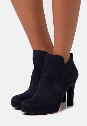 Tummansininen mokkanahkainen nilkkabootsi, jossa on tyylikäs muotoilu, korkea korko ja pyöristetty kärki. Täydentää aaltoileva yläreuna ja sileä pinta.