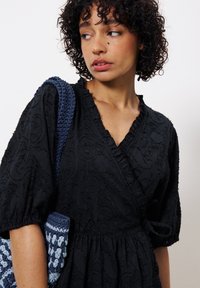 Schwarzes, strukturiertes Wickelkleid mit Puffärmeln und einem rüschierten Halsausschnitt, kombiniert mit einer blauen gehäkelten Tasche mit geometrischem Design.