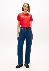 T-shirt rossa a maniche corte con la scritta "TOMMY HILFIGER EST. 1985", abbinata a jeans di denim blu a vita alta e mocassini neri. Look semplice e casual.