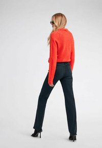 Maglione rosso peloso, jeans scuri, scarpe con tacco; persona di spalle, capelli lunghi, occhiali da sole, silhouette slanciata, dettagli minimali.