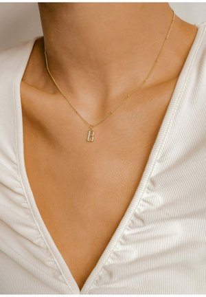 Goldkette mit einem diamantbesetzten Anhänger in Form des Buchstabens "B", zartes Kettenmuster und kleine Perlendetails. Im Hintergrund ist ein strukturierter weißer Stoff zu sehen.