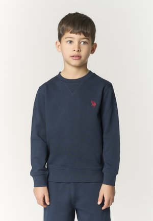 Ragazzo con capelli castani corti che indossa una felpa blu navy con logo rosso, in piedi contro uno sfondo chiaro e uniforme, che guarda leggermente a destra.