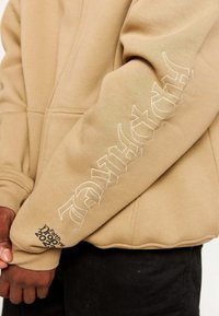 Felpa beige con cappuccio, caratterizzata da testo ricamato bianco sul maniche sinistra e dettaglio del logo vicino al polsino, realizzata in tessuto morbido con una vestibilità comoda.