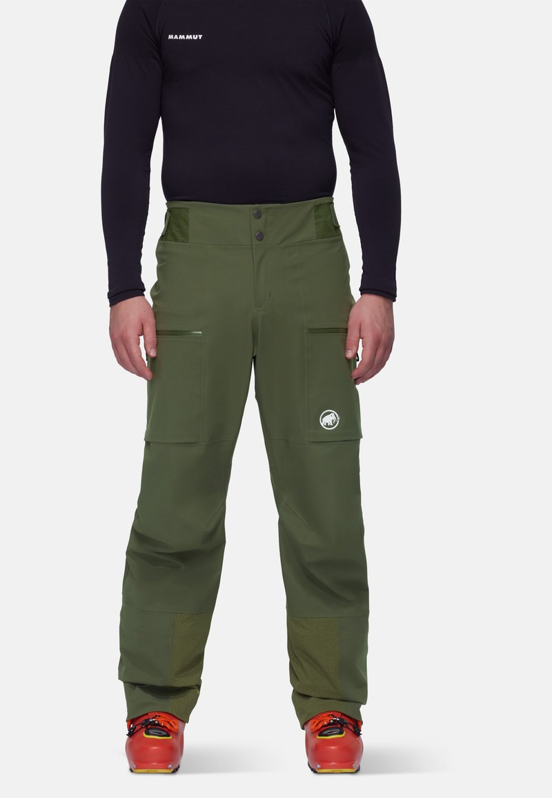Pantalons verts d'extérieur au design épuré, avec des poches zippées et des panneaux en mesh aux poignets. Assortis à des chaussures de ski rouge vif.
