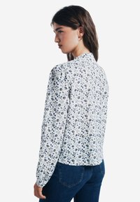 Bluse mit Blumenmuster aus weißem Stoff, mit blauen und grauen Blumenmustern, langen Ärmeln und dezenten Falten an den Schultern.