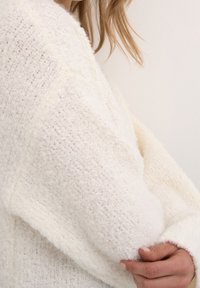 Pull blanc texturé à coupe ample, avec un tissu doux et moelleux et des manches larges, photographié de profil.