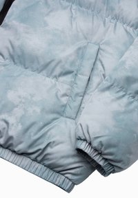 Hellblaue Daunenjacke mit einer glatten, strukturierten Oberfläche, elastischen Bündchen und einer Seitentasche. Der Stoff hat ein verblasstes Muster.
