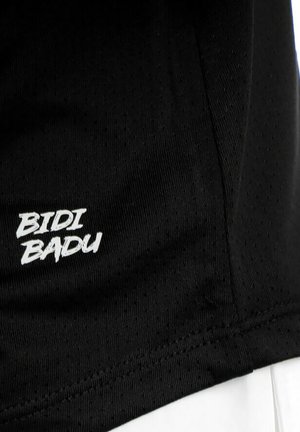 Tela negra con texto bordado en blanco "BIDI BADU" cerca de una costura, posiblemente parte de una prenda deportiva o ropa de entrenamiento.