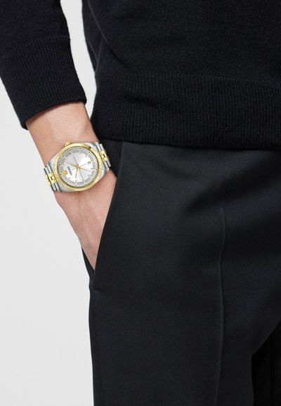 Main gauche dans la poche d'un pantalon noir, portant une montre-bracelet en argent et or avec un cadran rond blanc et une lunette ornée, pull noir à manches longues.