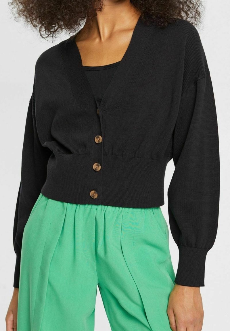 Cardigan nero lavorato a maglia con scollatura a V profonda, design corto, frontale con bottoni e maniche a sbuffo, abbinato a pantaloni plissettati verde menta.