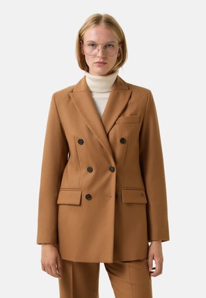 Femme avec des cheveux blonds courts portant de grandes lunettes transparentes, un blazer camel croisé, un pantalon assorti et un col roulé blanc.