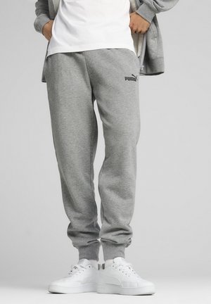 Person iført grå Puma joggingbukser, en hvid T-shirt, en grå hættetrøje med lynlås og hvide sneakers, stående med hænderne i lommerne.