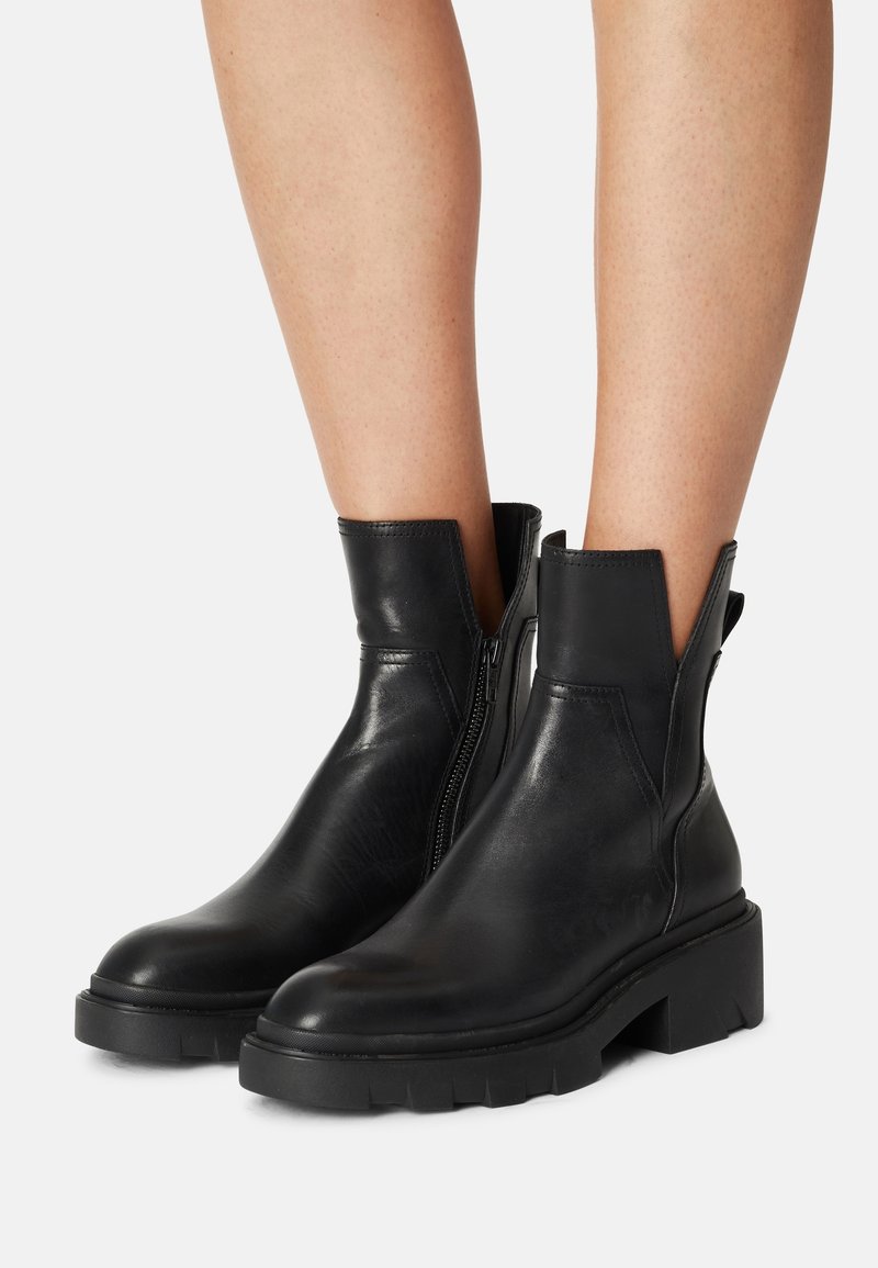 Bottes basses en cuir noir avec semelles épaisses et fermetures éclair latérales portées sur jambes nues sur fond uni.
