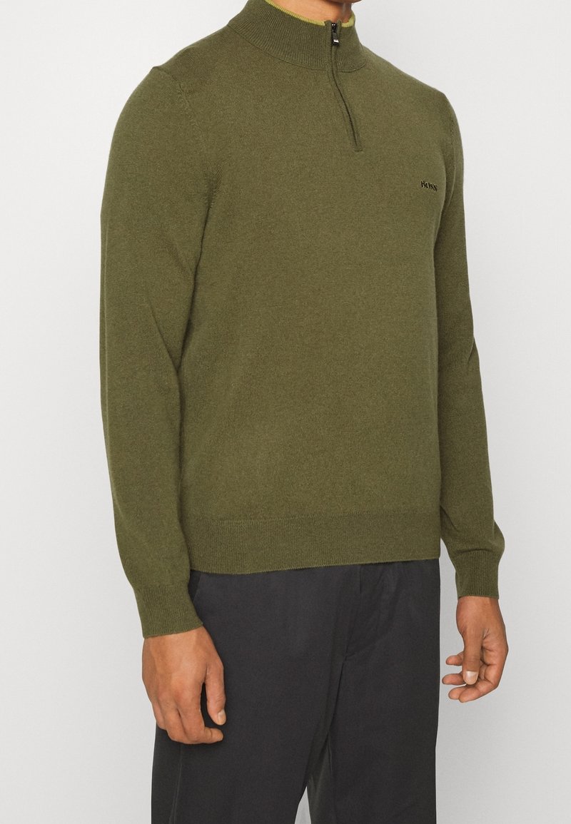 Mies, joka pukeutuu oliivinvihreään pitkähihaiseen puoleen vetoketjulliseen neuleeseen, jonka hihansuissa ja kauluksessa on ribbineule, yhdistettynä mustiin housuihin, yksinkertaista valkoista taustaa vasten.