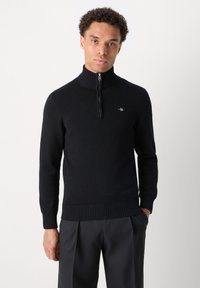 Joven con un jersey negro de canalé con cremallera en el cuello y pantalones oscuros, de pie con una mano en el bolsillo contra un fondo liso.