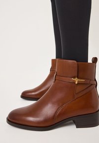 Bottines en cuir marron avec un léger talon, dotées d'une accentuation dorée et d'un design épuré et minimaliste. Fini texturé sur le dessus.