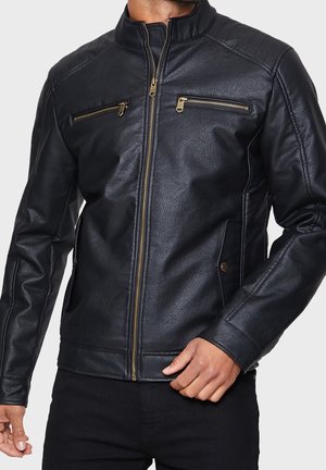 Hombre con chaqueta de cuero negra texturizada con cremalleras y bolsillos dorados, ajustando el dobladillo inferior mientras está de pie.