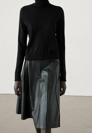 Personne portant un pull à col roulé noir et une jupe en cuir noire longueur genou, debout devant un fond uni.