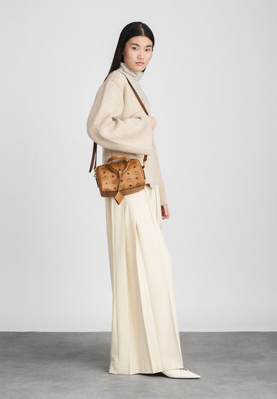 Kvinde iført en beige cardigan, stribet turtleneck, brede cremefarvede bukser, hvide sko, bærende en brun designer skuldertaske med logoer.