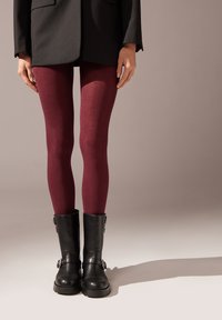 Collant bordeaux con una texture morbida, abbinati a stivali neri alla caviglia con fibbie, su uno sfondo neutro.