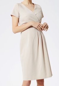 Vestido envolvente beige con mangas cortas, escote en V y falda plisada. Elaborado con una tela suave, presenta un corsé ajustado y un diseño fluido.