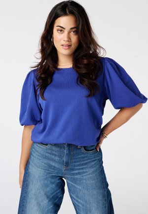 Vrouw met lang donker haar die een blauw top met pofmouwen en een blauwe spijkerbroek draagt, staand met één hand op haar heup tegen een effen achtergrond.