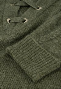 Close-up van een olijfgroen gebreide trui met geribde boord en sluitingen met knoppen op een gestructureerde stof.