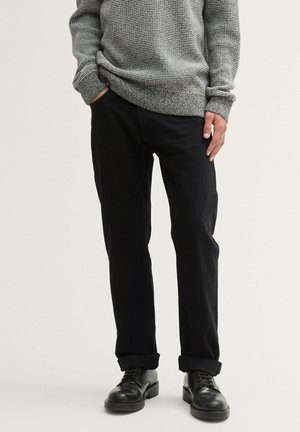 Schwarze Jeans mit geradem Schnitt, getragen mit einem umgeschlagenen Saum, kombiniert mit einem strukturierten grauen Langarmpullover und schwarzen Schnürschuhen.