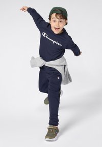 Ragazzo che indossa un tuta Champion blu scuro, un cappellino verde e scarpe da ginnastica, che corre gioiosamente con le braccia aperte su sfondo bianco.