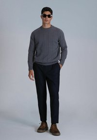Pull en maille gris avec des rayures verticales, associé à un pantalon bleu marine. Des chaussures à lacets marron complètent le look. Le modèle porte des lunettes de soleil noires.