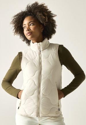 Femme aux cheveux bouclés portant une veste matelassée légère sur une chemise à manches longues couleur vert olive, debout les mains dans les poches de la veste.