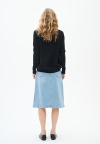 Pull en tricot noir, jupe en denim bleu avec deux poches arrière, longueur sous le genou, portée avec des chaussures plates noires, vue de dos.
