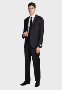 Completo nero con revers in raso, camicia bianca, cravatta nera e scarpe nere lucide. Include un fazzoletto da taschino e dettagli di cucitura sottili.