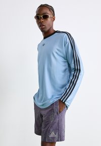 Nuori mies, jolla on päällään vaaleansininen Adidasin pitkähiha paita mustilla raidoilla ja violetit Adidasin shortsit, seisoo kädet taskuissa aurinkolasit päässään.