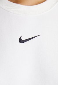 Λευκή αθλητική μπλούζα από μαλακό ύφασμα με υφή, διακοσμημένη με ένα μικρό μαύρο κεντημένο λογότυπο Nike στο στήθος.