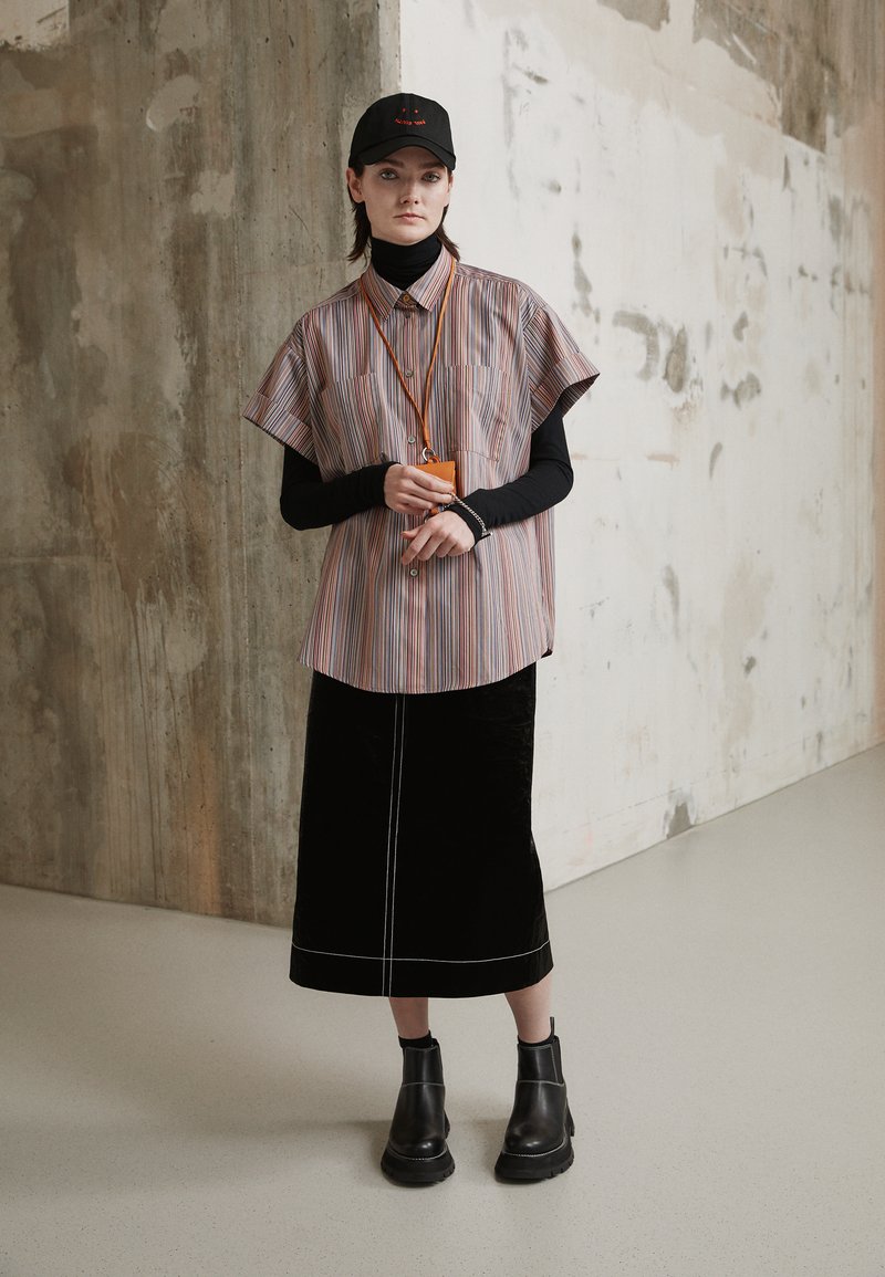 Camisa de manga curta às riscas com colarinho, suéter de gola alta preto por baixo, saia longa de veludo preto e botas de cano curto pretas. Acess�ório laranja ao redor do pescoço.