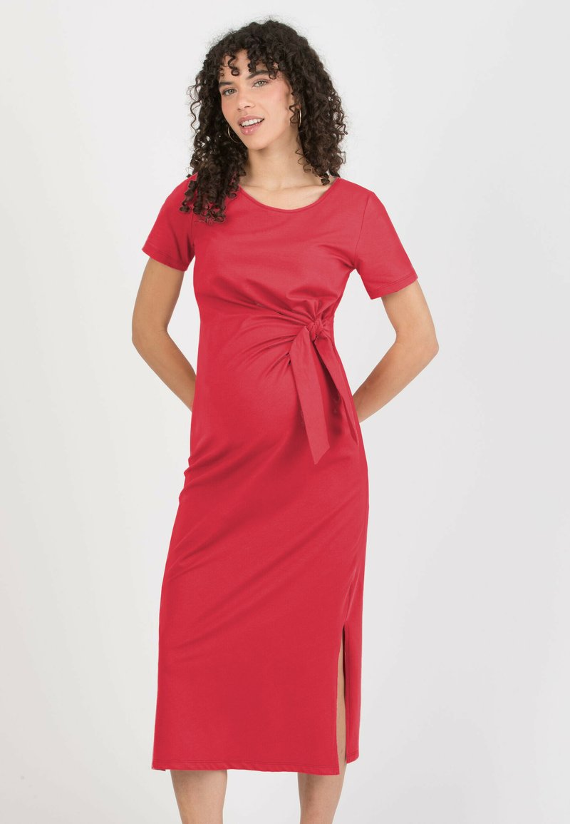 Robe rouge mi-longue en tissu lisse. Elle présente des manches courtes, un détail torsadé à l'avant avec un nœud, et une fente latérale pour faciliter les mouvements.