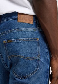 Jeans de denim en azul con un acabado texturizado, que presentan una etiqueta de cuero marrón en la cinturilla y costuras decorativas en los bolsillos traseros.